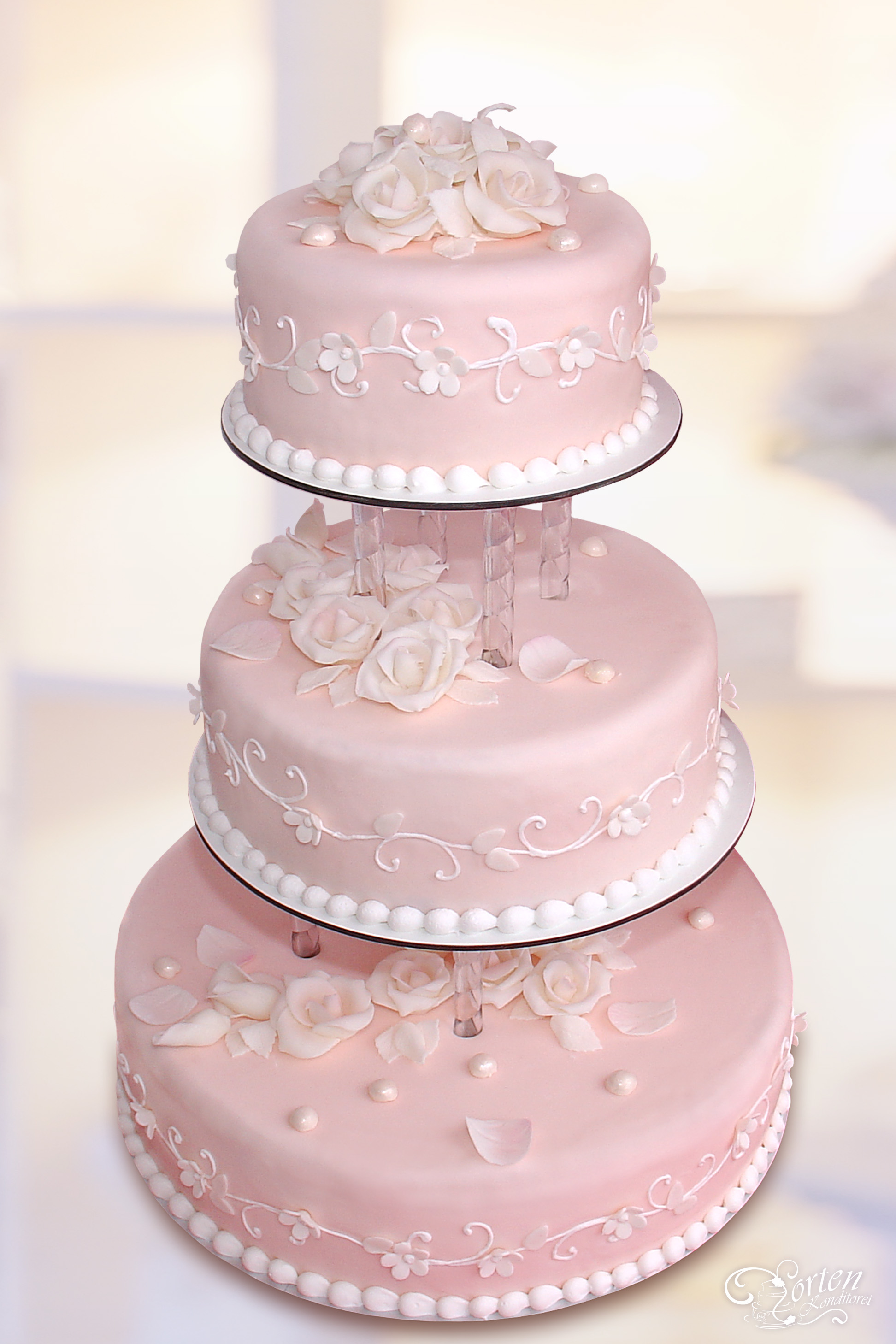 Für Romantiker: Dreistöckige Torte in pastell-rosa mit weißen Blüten und Schnörkeln sowie weißen Rosen und Blütenblättern.